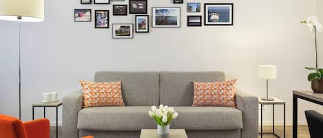 A modern living room with a grey sofa, orange pillows, a small table with a vase of flowers, and a wall decorated with framed pictures.