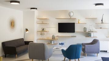 A modern living room with a flat-screen TV, grey and blue armchairs, a dark sofa, and wooden shelves with various decorative items.