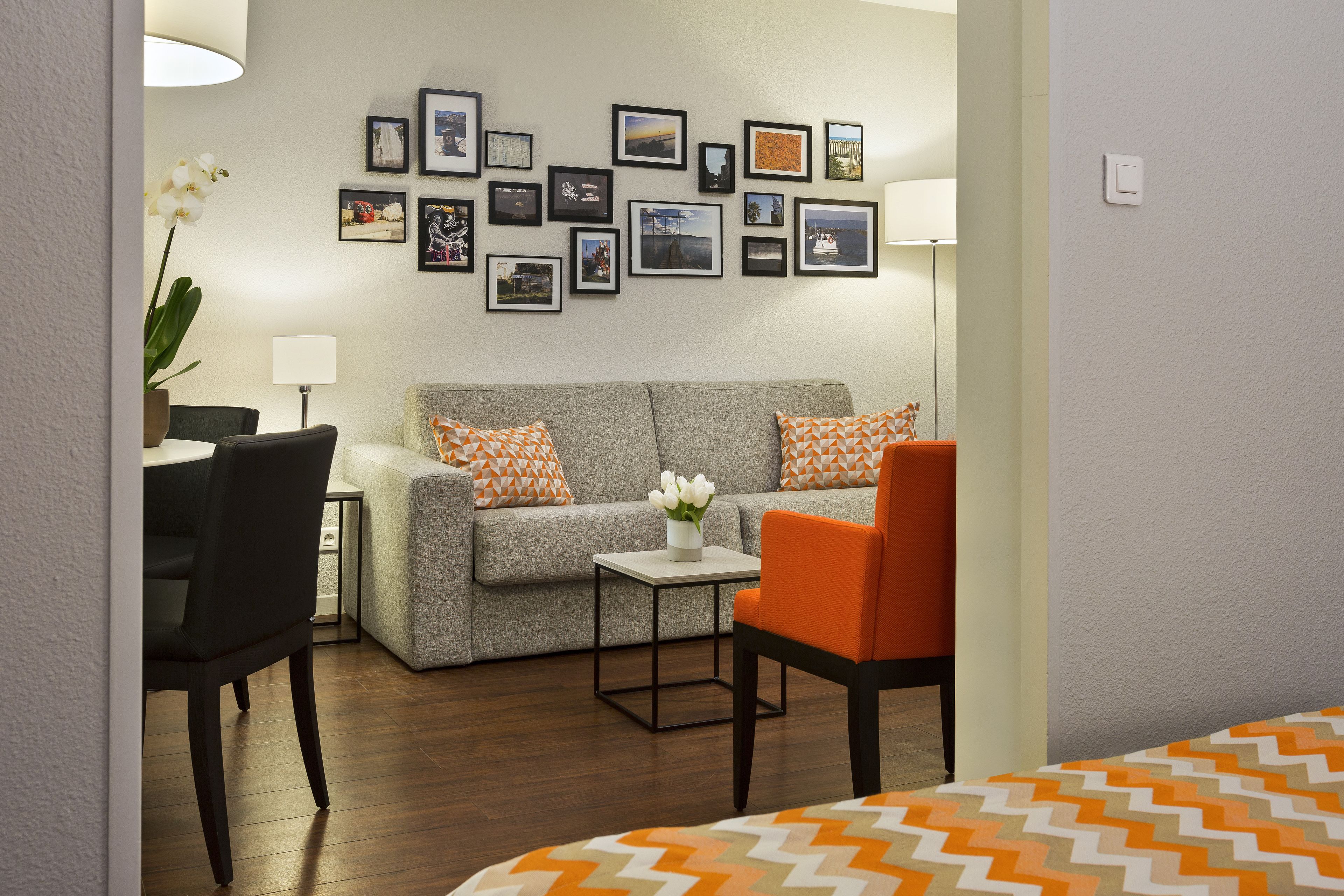 A modern living room with a grey sofa, orange and white patterned pillows, a small table with a vase of white flowers, and a black chair.