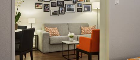 A modern living room with a grey sofa, orange and white patterned pillows, a small table with a vase of white flowers, and a black chair.