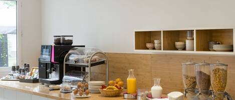 Buffet de café da manhã com máquinas de café, frutas, cereais e dispensadores de leite.