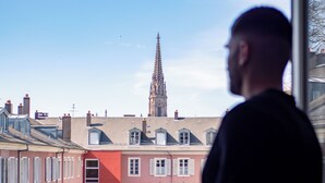 Una persona de pie junto a una ventana con vistas a un paisaje urbano, destacando una aguja de iglesia.