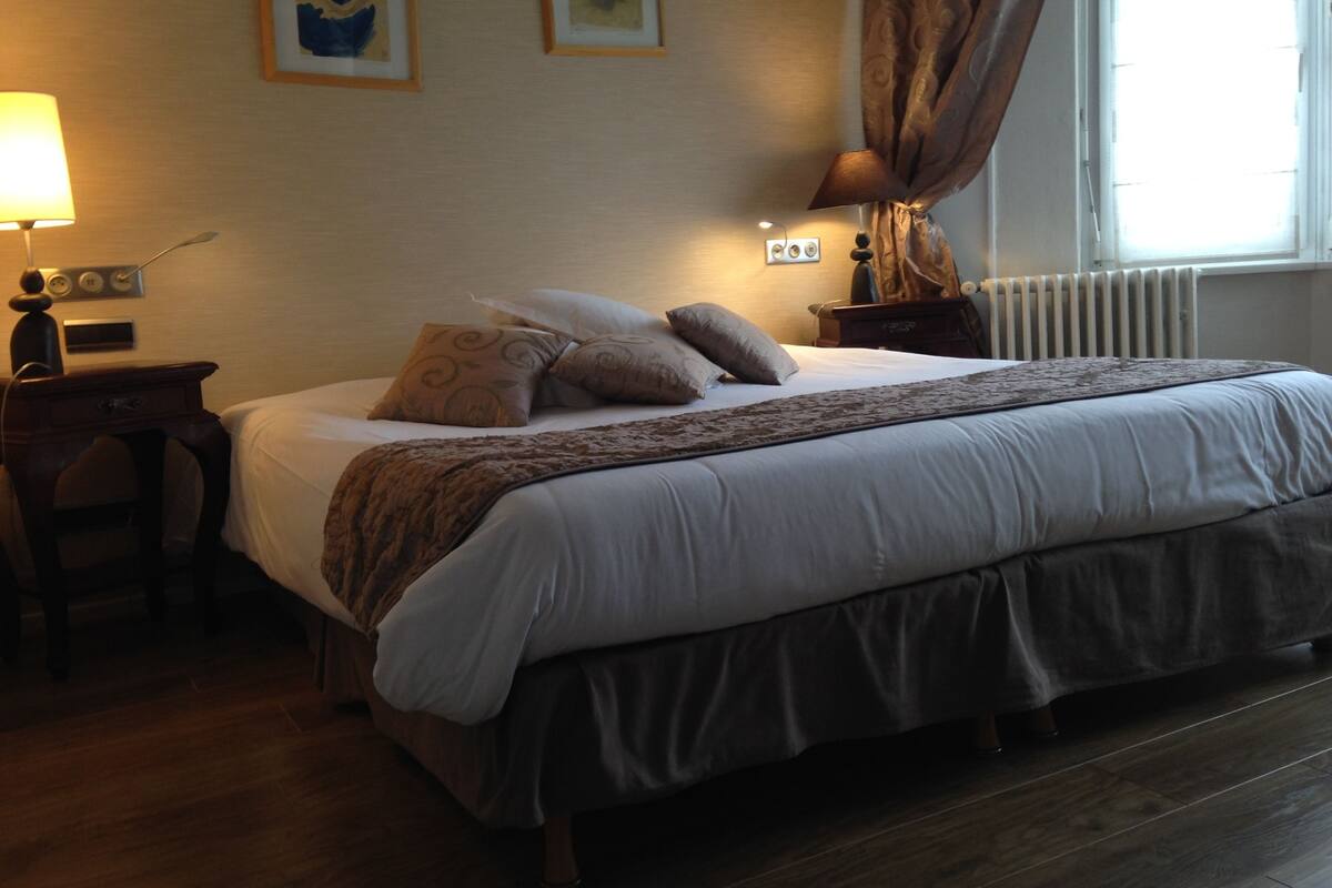 Un lit bien fait, recouvert d’un couvre-lit à motifs, une table de chevet avec une lampe, et deux œuvres d’art encadrées accrochées au mur.