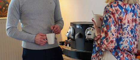 A man and a woman standing in a room with a coffee machine and a lamp in the background.