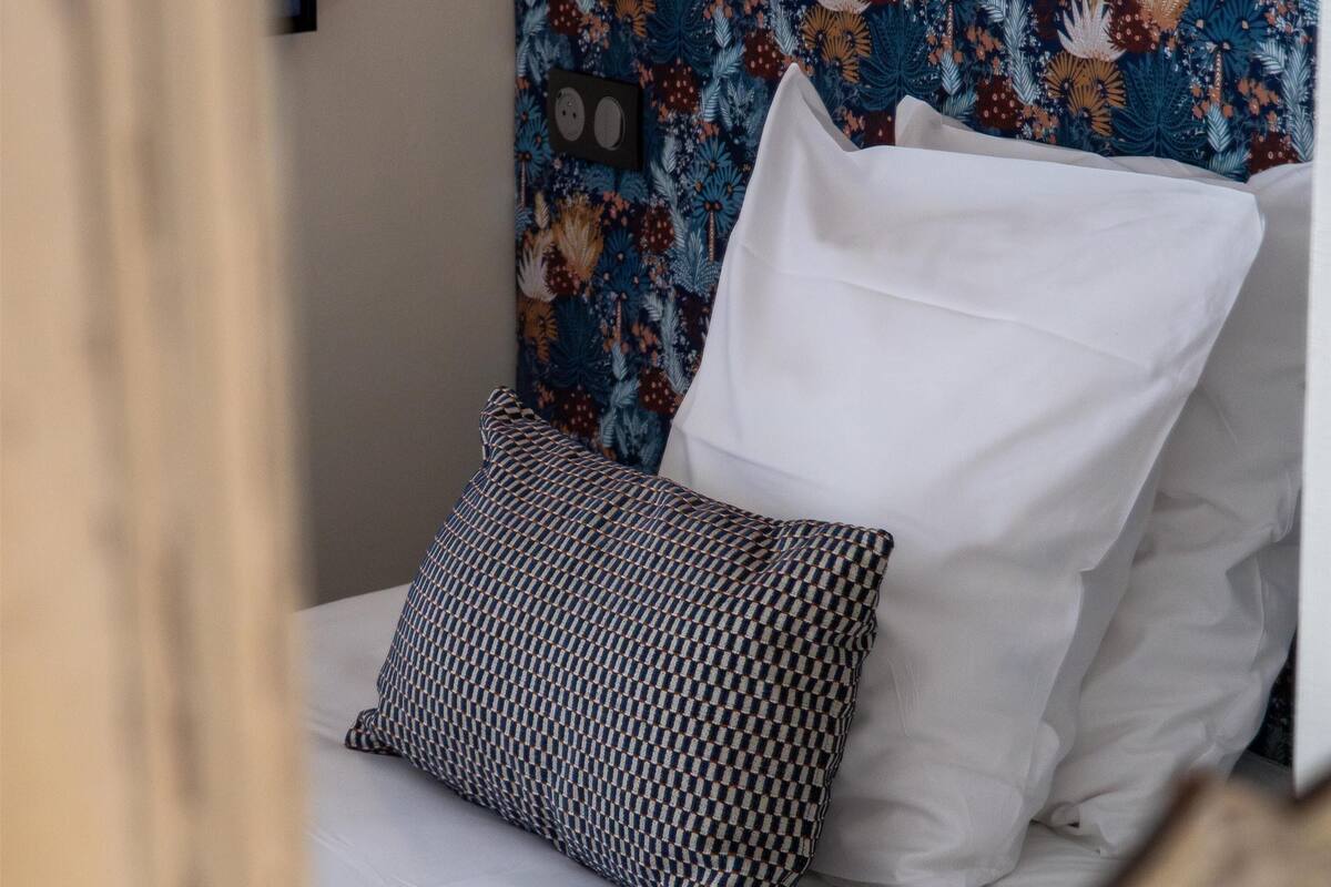 Un lit avec des draps blancs et des coussins à motifs, placé devant une tête de lit ornée de motifs floraux.