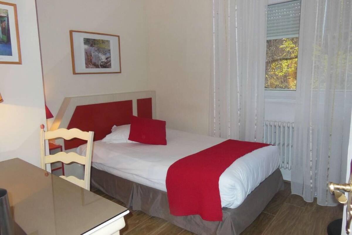 Un lit simple avec une tête de lit rouge, une table de chevet, une chaise et une fenêtre avec des rideaux.