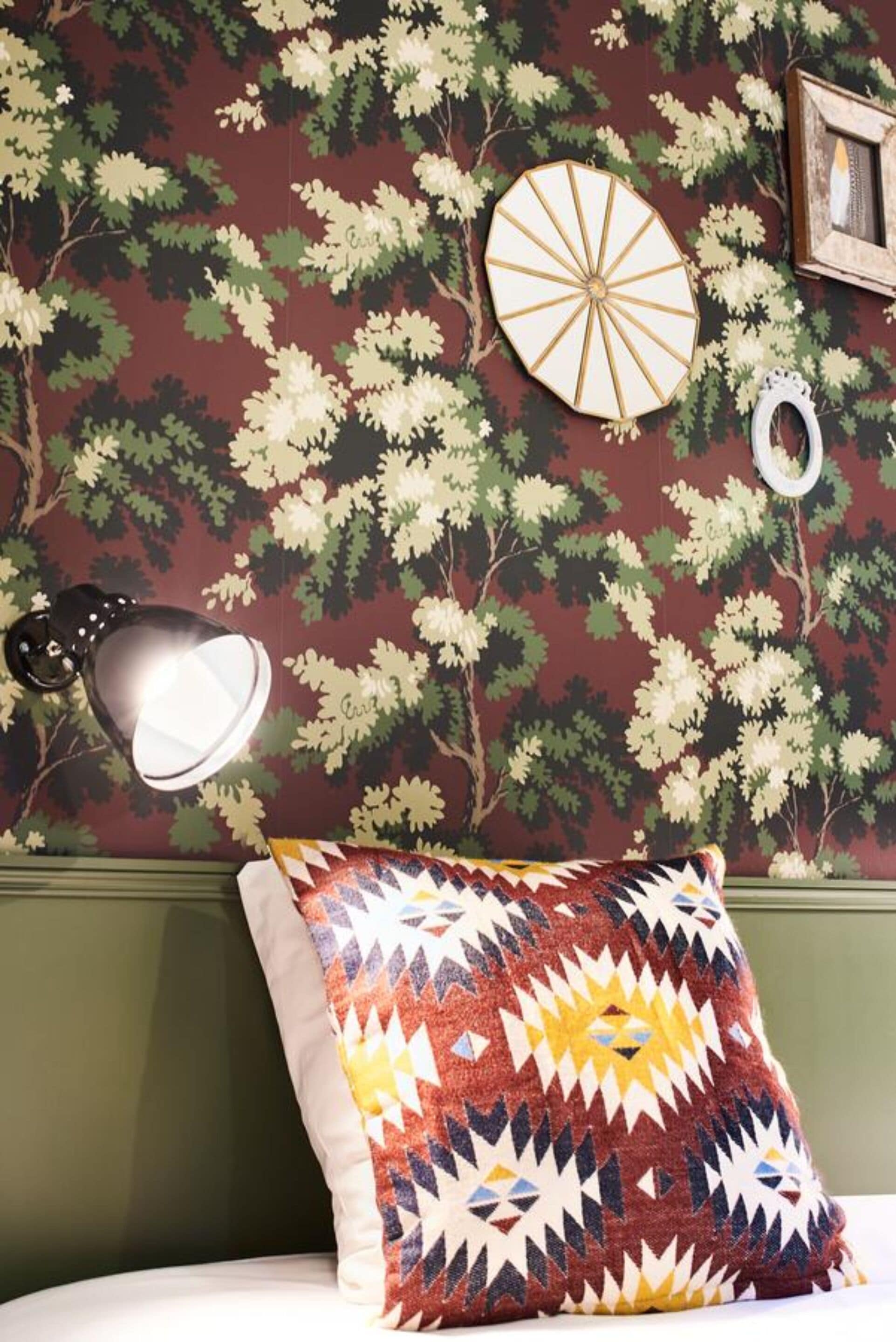 A room with a patterned wallpaper, a round wall decoration, a framed picture, and a colorful pillow on a bed.