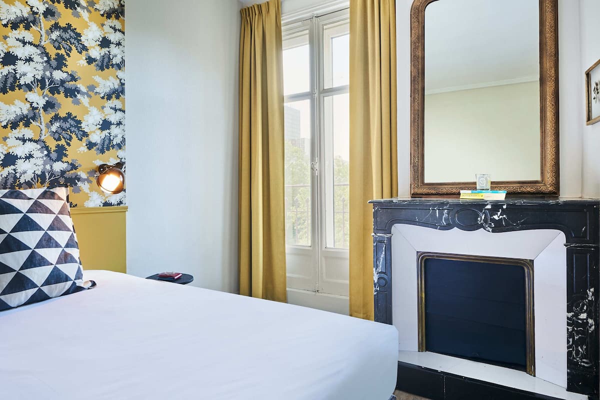 Habitación de hotel con cama, mesita de noche, espejo, chimenea y ventana con cortinas.
