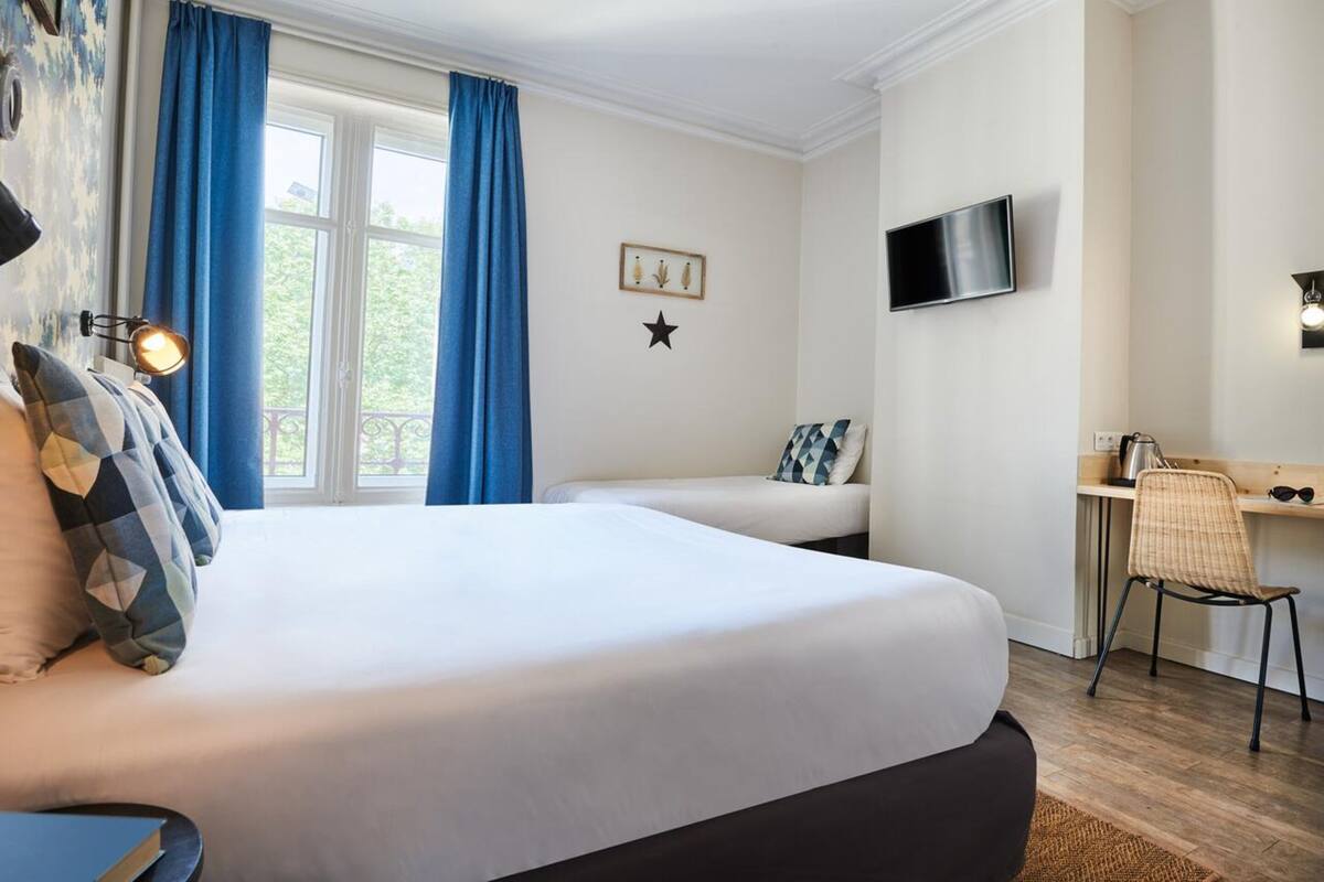 Un dormitorio con una cama grande, una cama más pequeña, un escritorio con silla, un televisor y una ventana con cortinas.