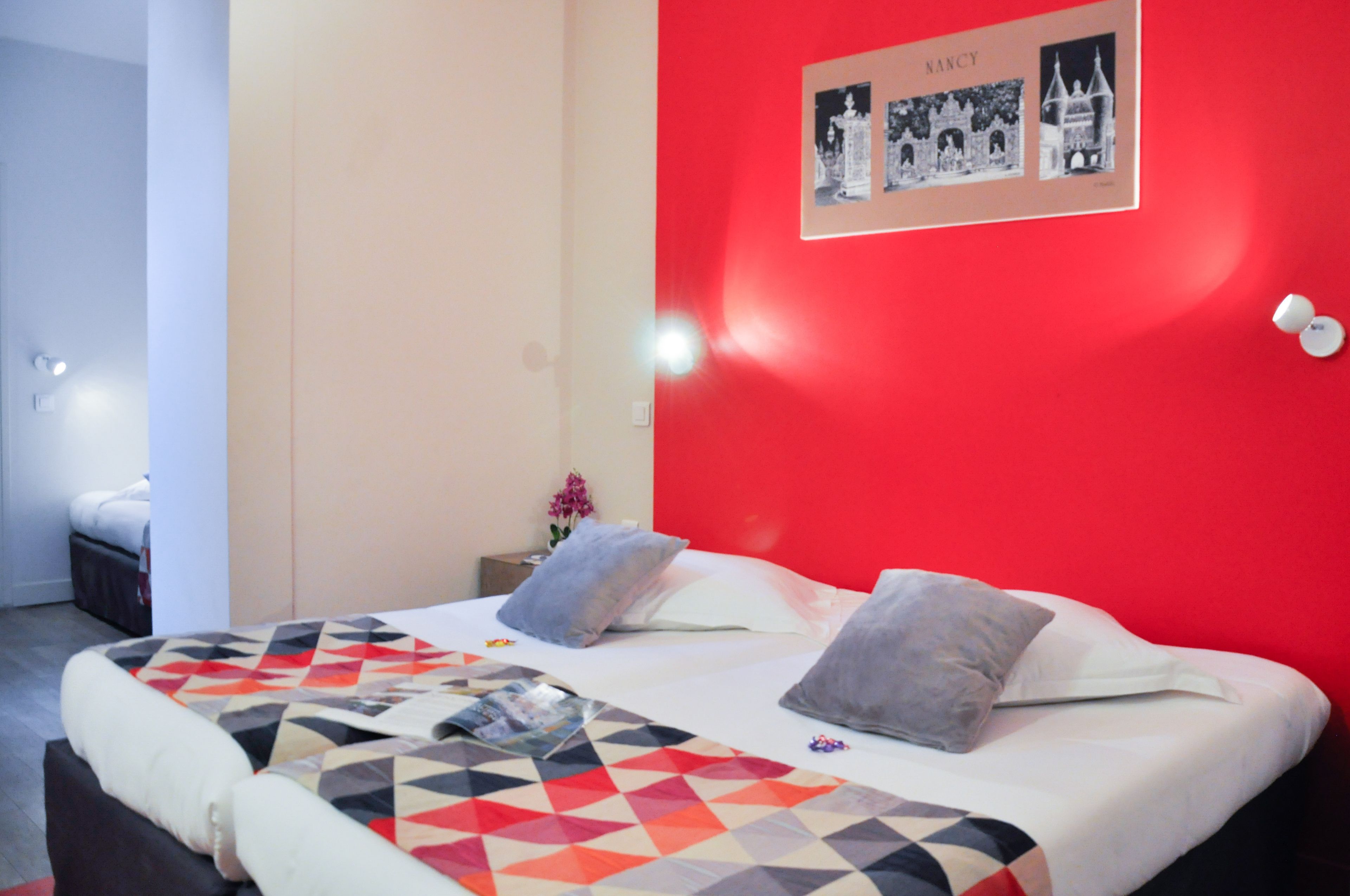 Un dormitorio con una pared de acento roja, una cama con colcha de diseño geométrico y un marco de pared con el nombre "NANCY".