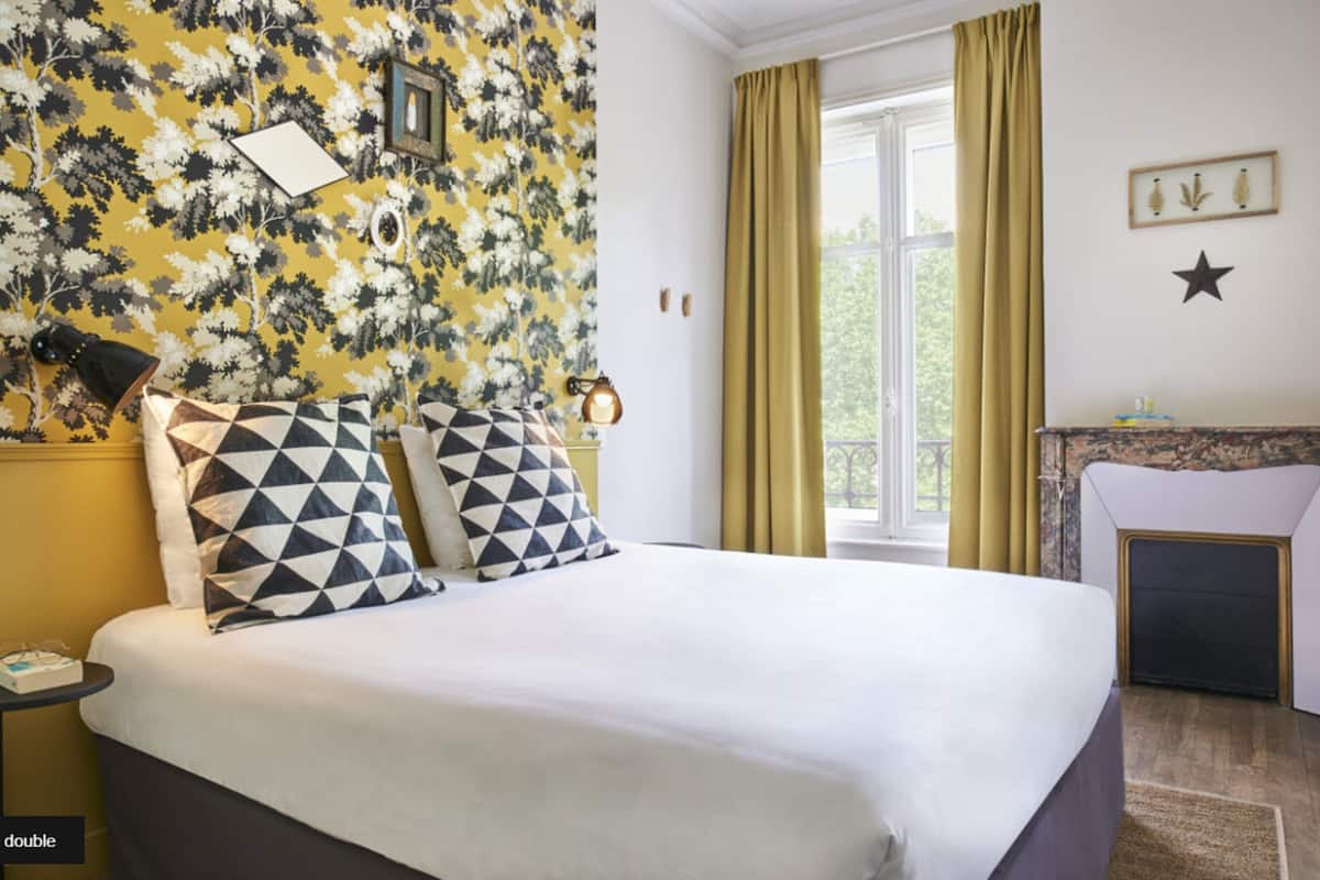 Habitación de hotel con cama, mesita de noche, lámpara, ventana con cortinas y chimenea.
