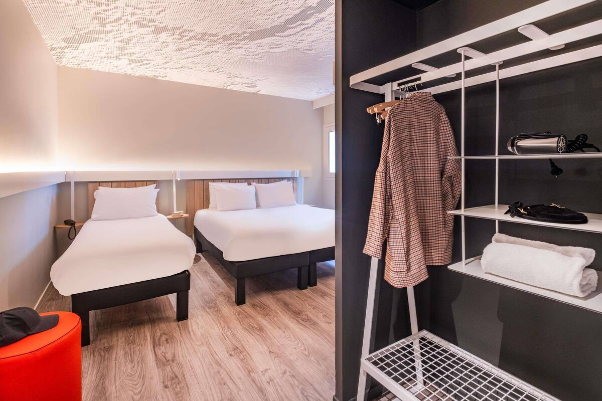 Une chambre d’hôtel avec deux lits, un pouf rouge, un porte-serviettes blanc et une étagère à chaussures.