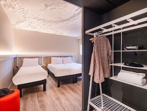 A hotel room with two beds, a red ottoman, a white towel rack, and a shoe shelf.