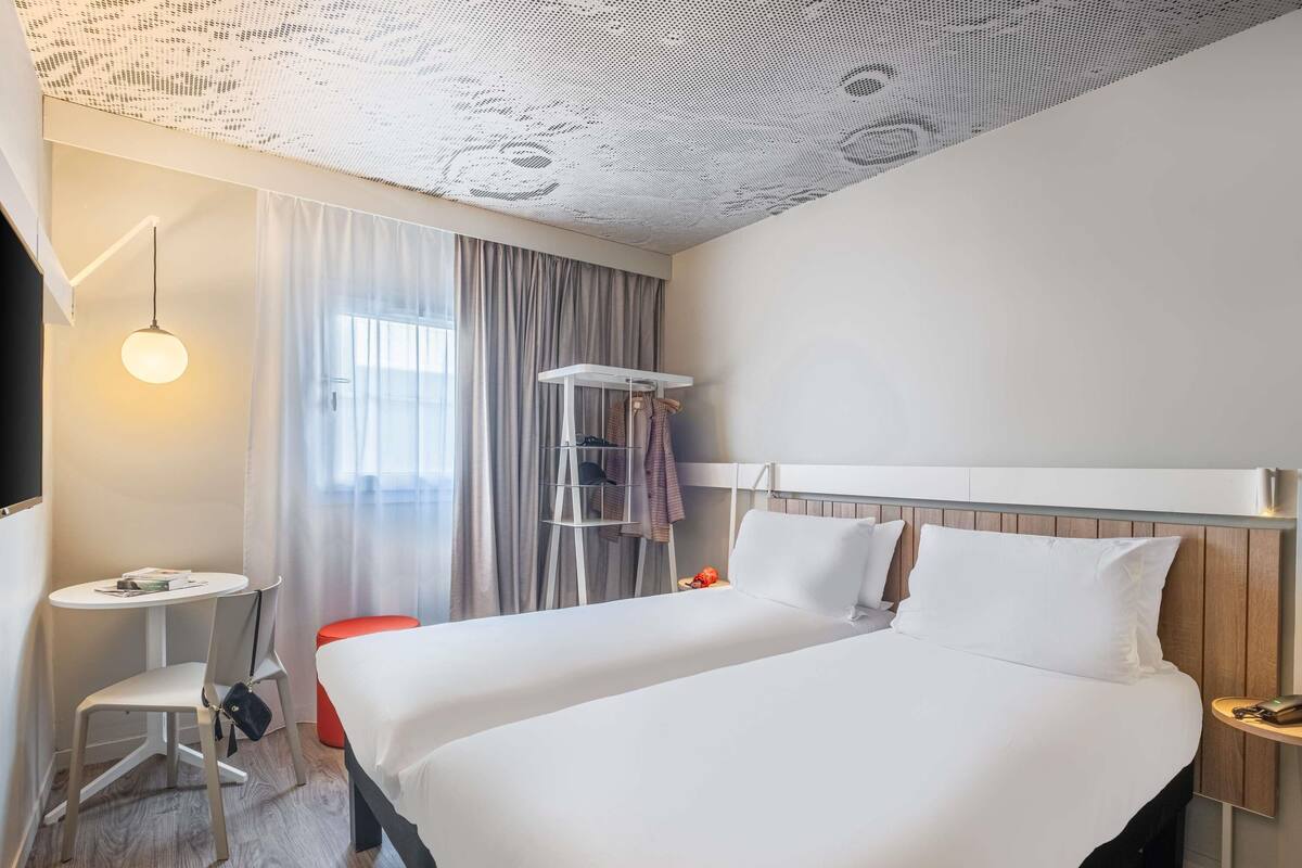 Une chambre d’hôtel avec un grand lit, un petit bureau, une chaise, un porte-manteau mural et un plafond orné d’un motif ajouré.