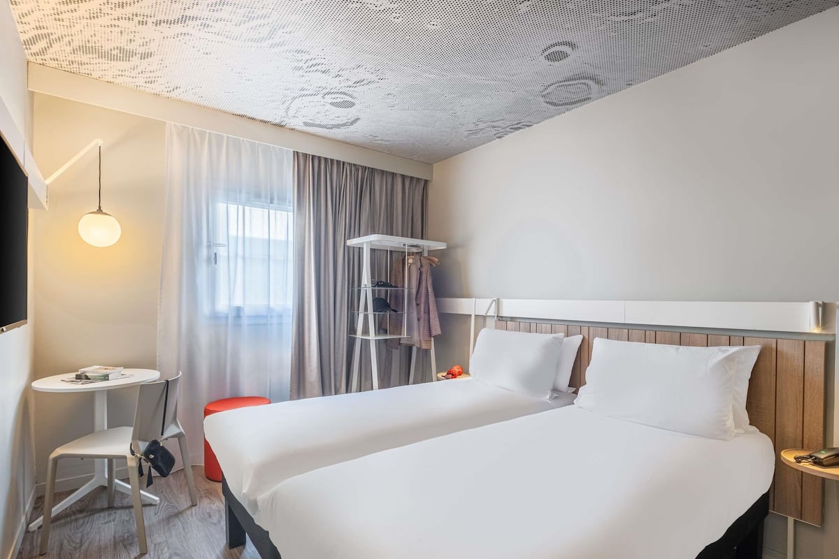 A hotel room with a large bed, a small desk, a chair, a hanging coat rack, and a ceiling with a perforated design.