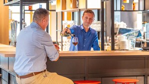 A bar area with a bartender pouring a drink. There are shelves with various bottles and glasses behind the bartender. - Ibis Nantes Nord Treillières (Treillieres)