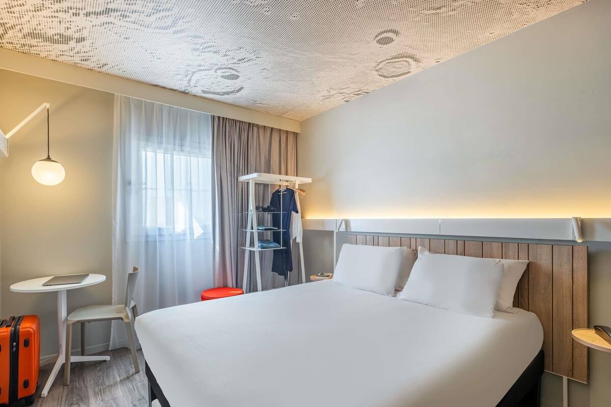 Une chambre d’hôtel moderne avec un grand lit, une petite table, une chaise, une valise et un porte-vêtements.