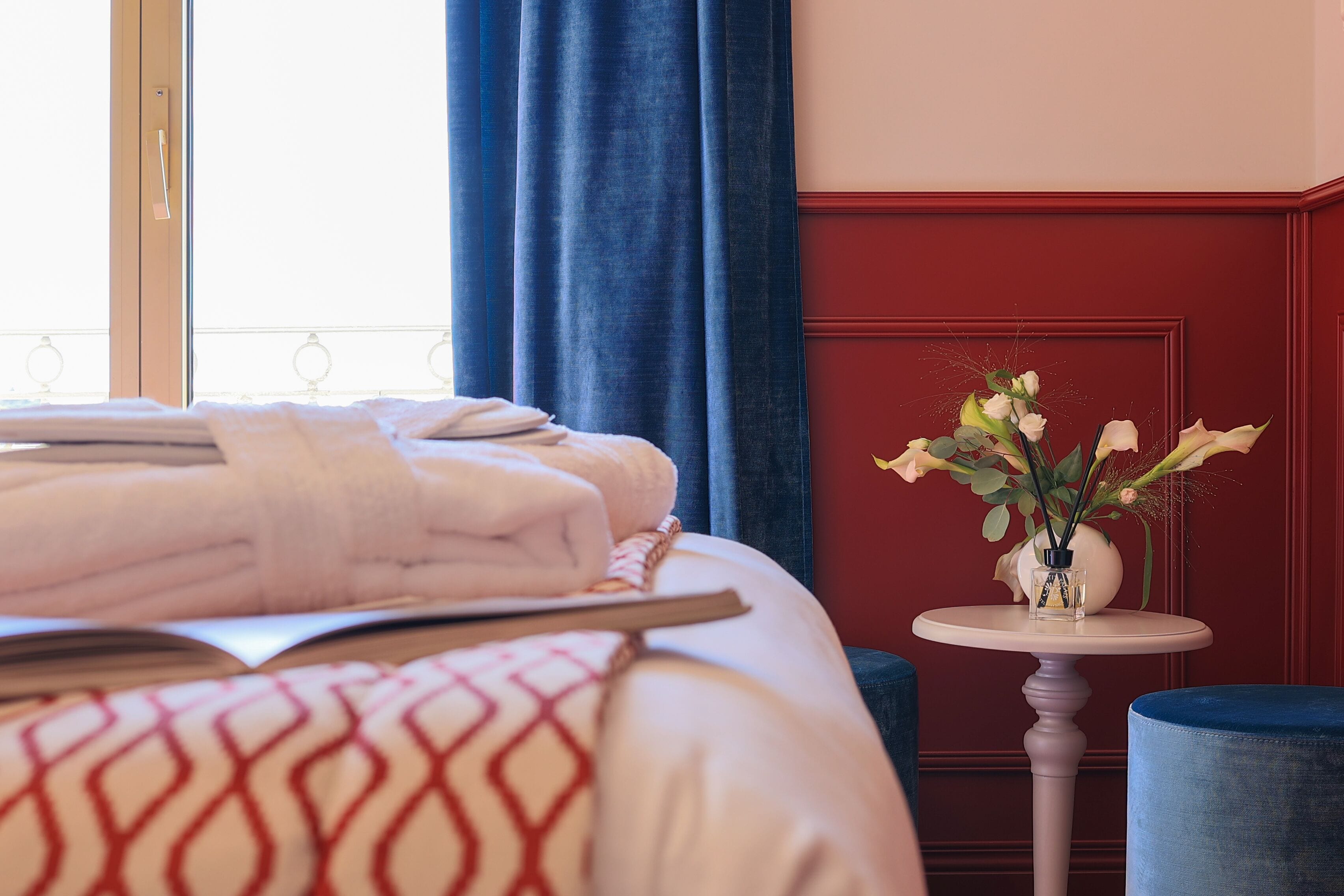 A neatly made bed with a patterned pillow, a bedside table with a vase of flowers, and a blue ottoman.