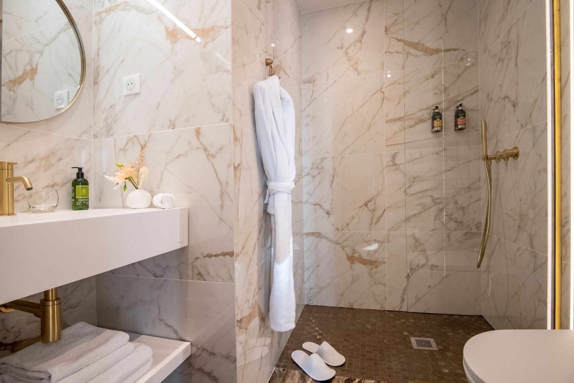 A modern bathroom with a marble shower, a vanity with a gold faucet, and toiletries.