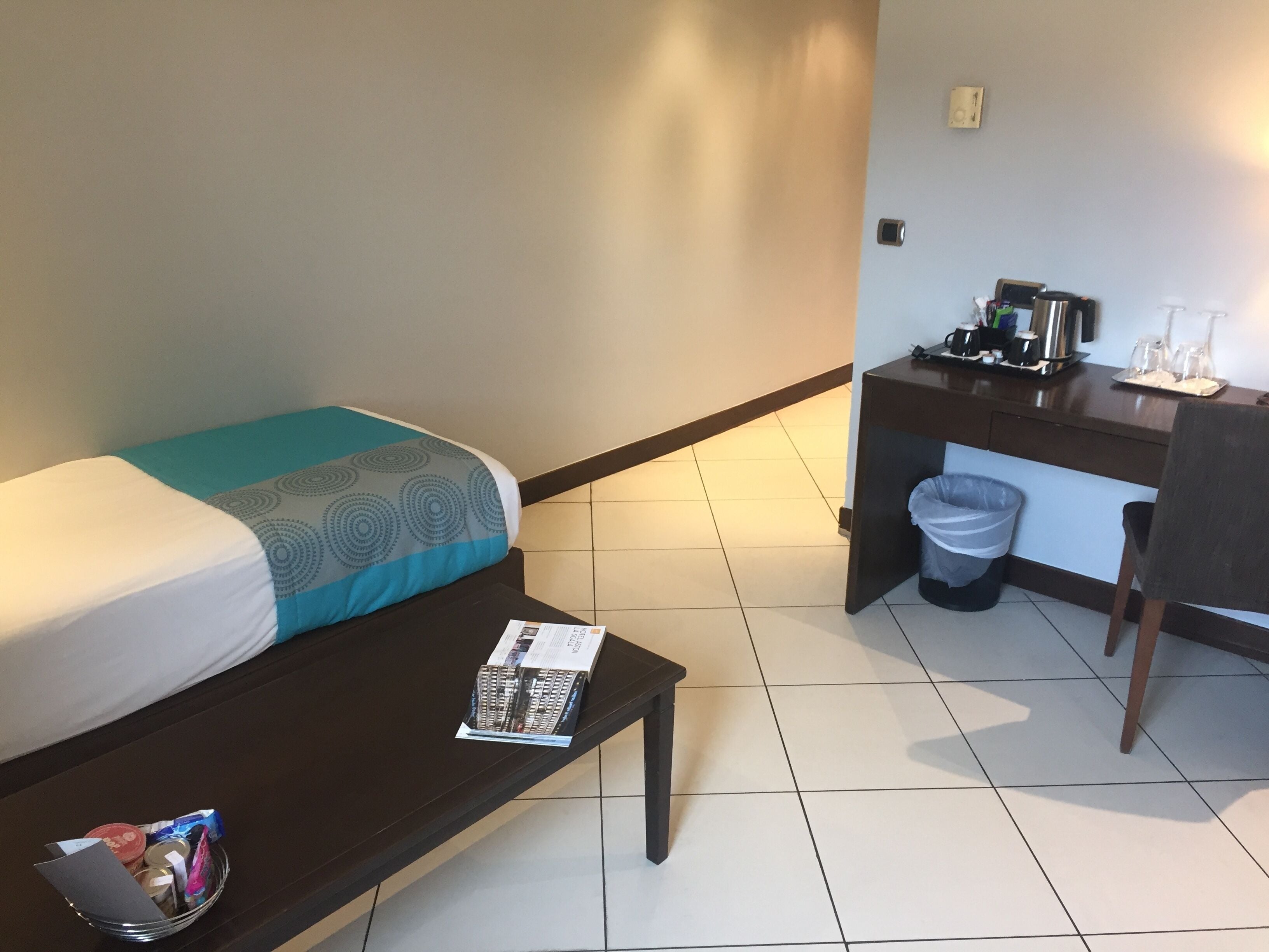 A hotel room with a bed, a small table with a magazine, a desk with a coffee maker, and a trash bin.