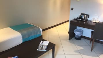 A hotel room with a bed, a small table with a magazine, a desk with a coffee maker, and a trash bin.