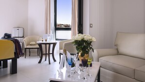 A hotel room with a sofa, a dining table with glasses, a vase with flowers, and a view of buildings through a window. - Hôtel Aston La Scala (Nice)