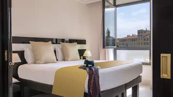 Habitación de hotel con una cama grande, una mesita de noche con lámpara y vistas a edificios a través de una ventana.