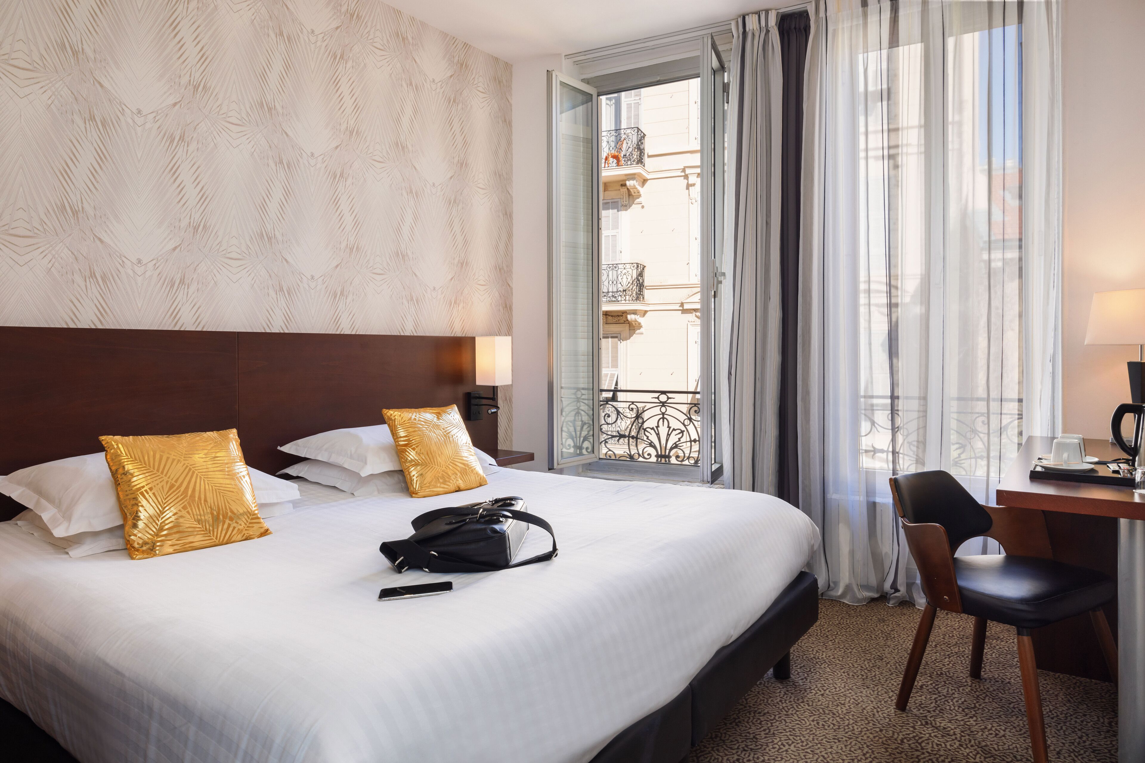 A hotel room with a large bed, two gold-patterned pillows, a black handbag, a desk with a chair, and a view of balconies through sheer curtains.