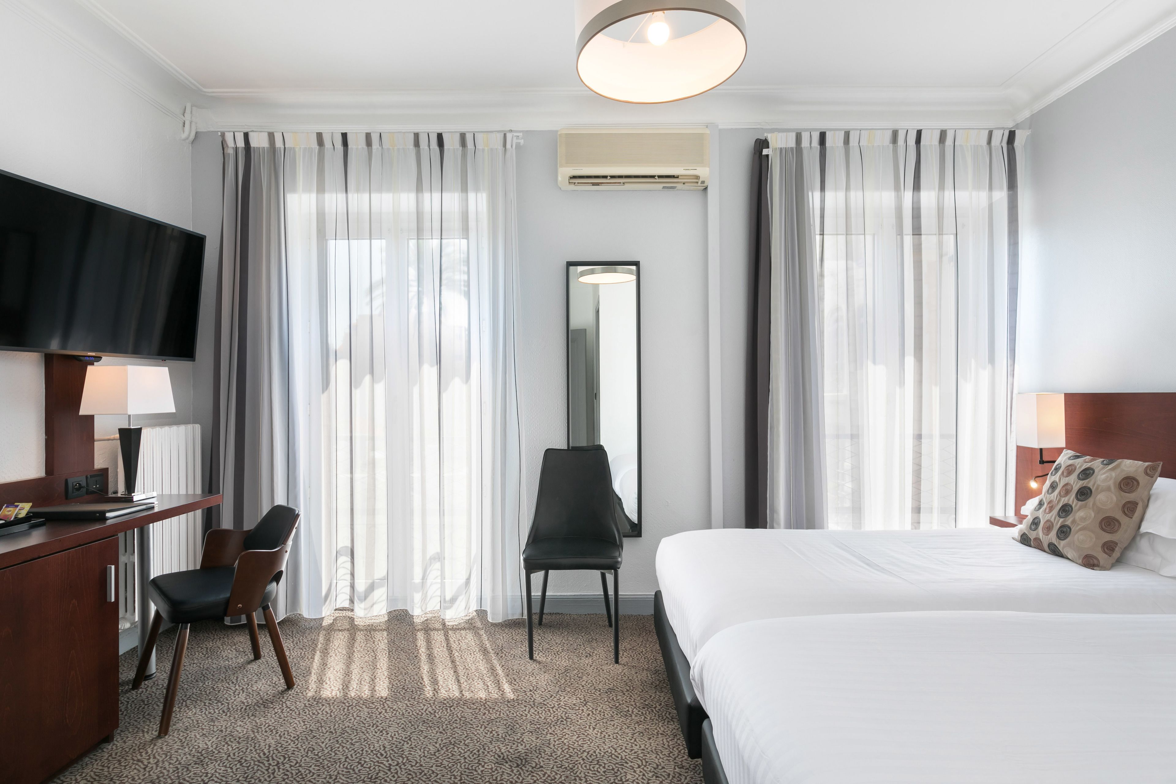 A hotel room with a bed, a desk with a chair, a TV, and a large window with curtains.