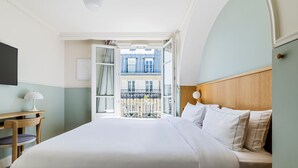 A hotel room with a bed, a desk, and a view of a building through an open door. - Hôtel Oratio (Paris)