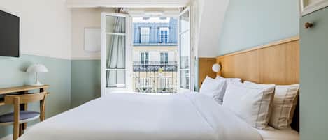 A hotel room with a bed, a desk, and a view of a building through an open door.