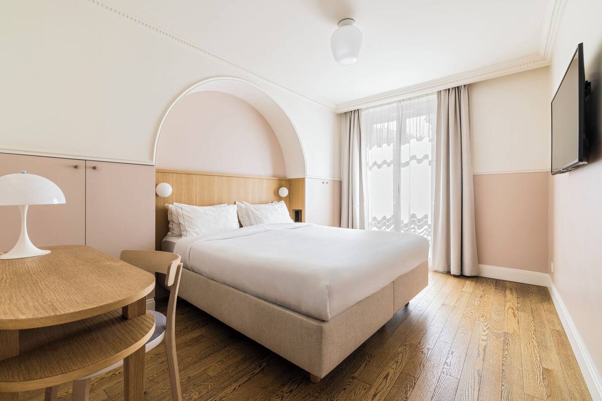 A hotel room with a bed, a round wooden table, a chair, a TV mounted on the wall, and a window with curtains.