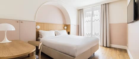 Habitación de hotel con cama, una mesa redonda de madera, una silla, televisor montado en la pared y una ventana con cortinas.