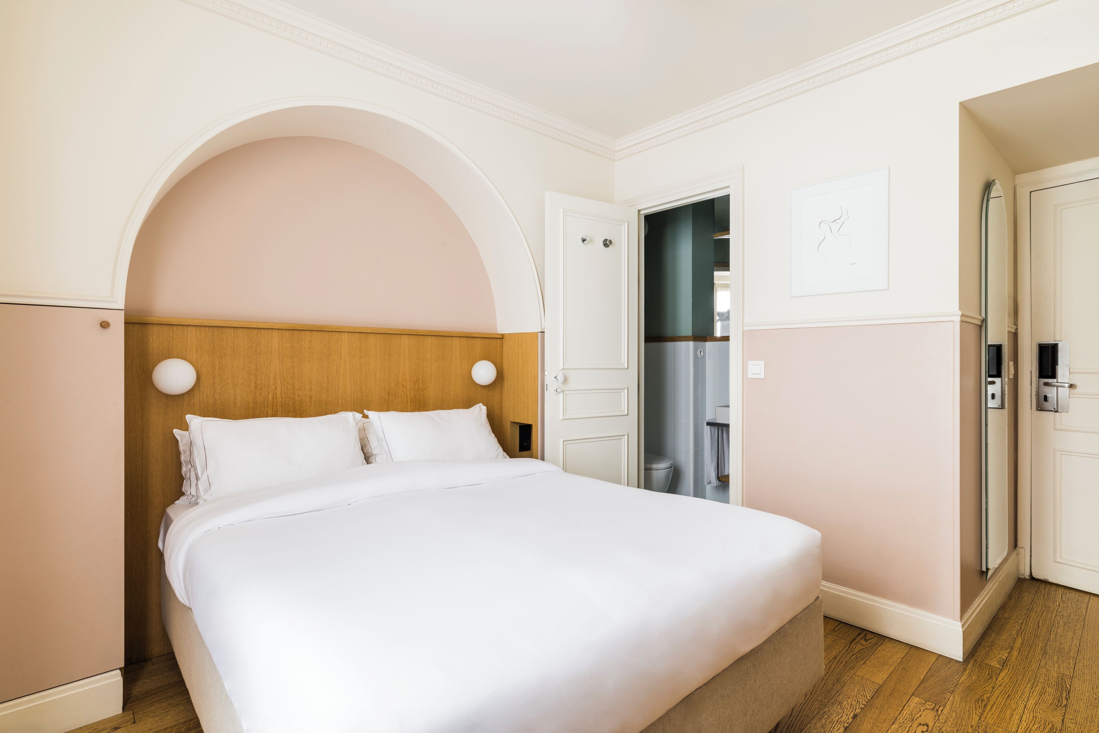 A hotel room with a large bed, wooden headboard, white walls, and a door leading to a bathroom.