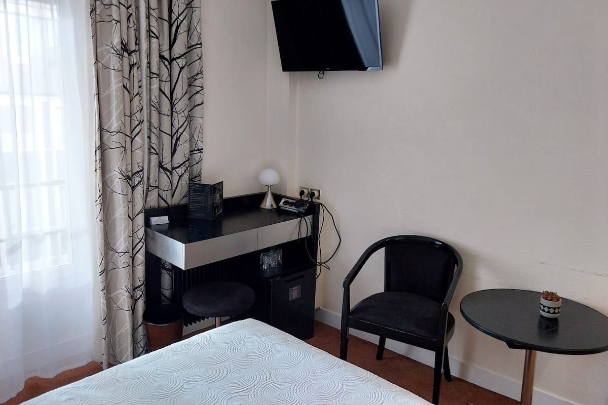 Una habitación de hotel con cama, escritorio y silla, televisor montado en la pared, una mesita y una ventana con cortinas translúcidas.