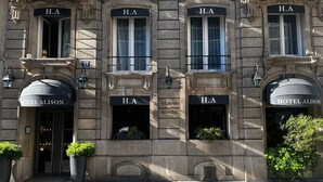 Un edificio con múltiples letreros H.A sobre ventanas y puertas, que indican habitaciones de hotel. Hay plantas en macetas en la entrada y una puerta de acceso visible con el rótulo "HOTEL ALISON".