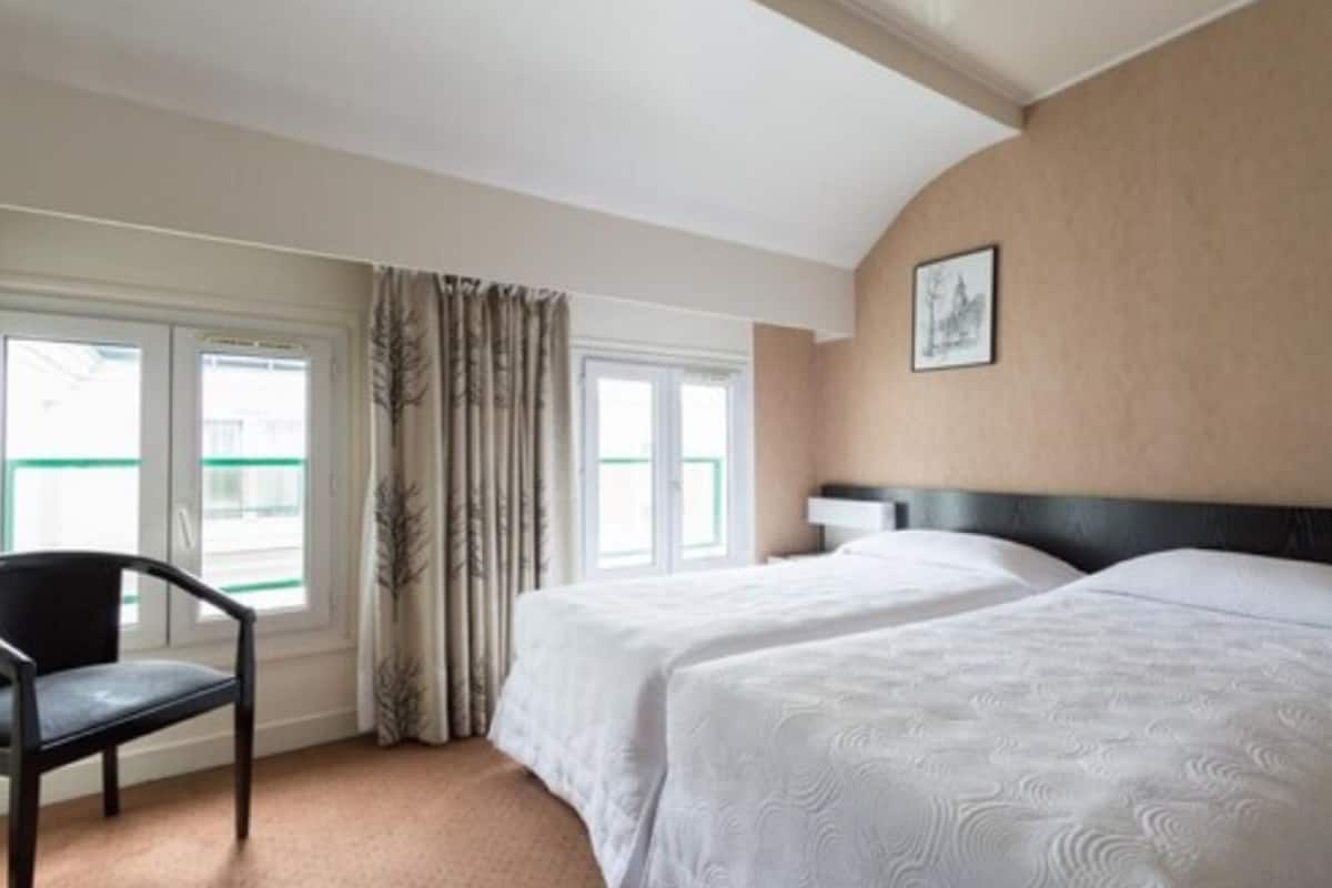 Habitación de hotel con dos camas, una silla y un ventanal con cortinas.