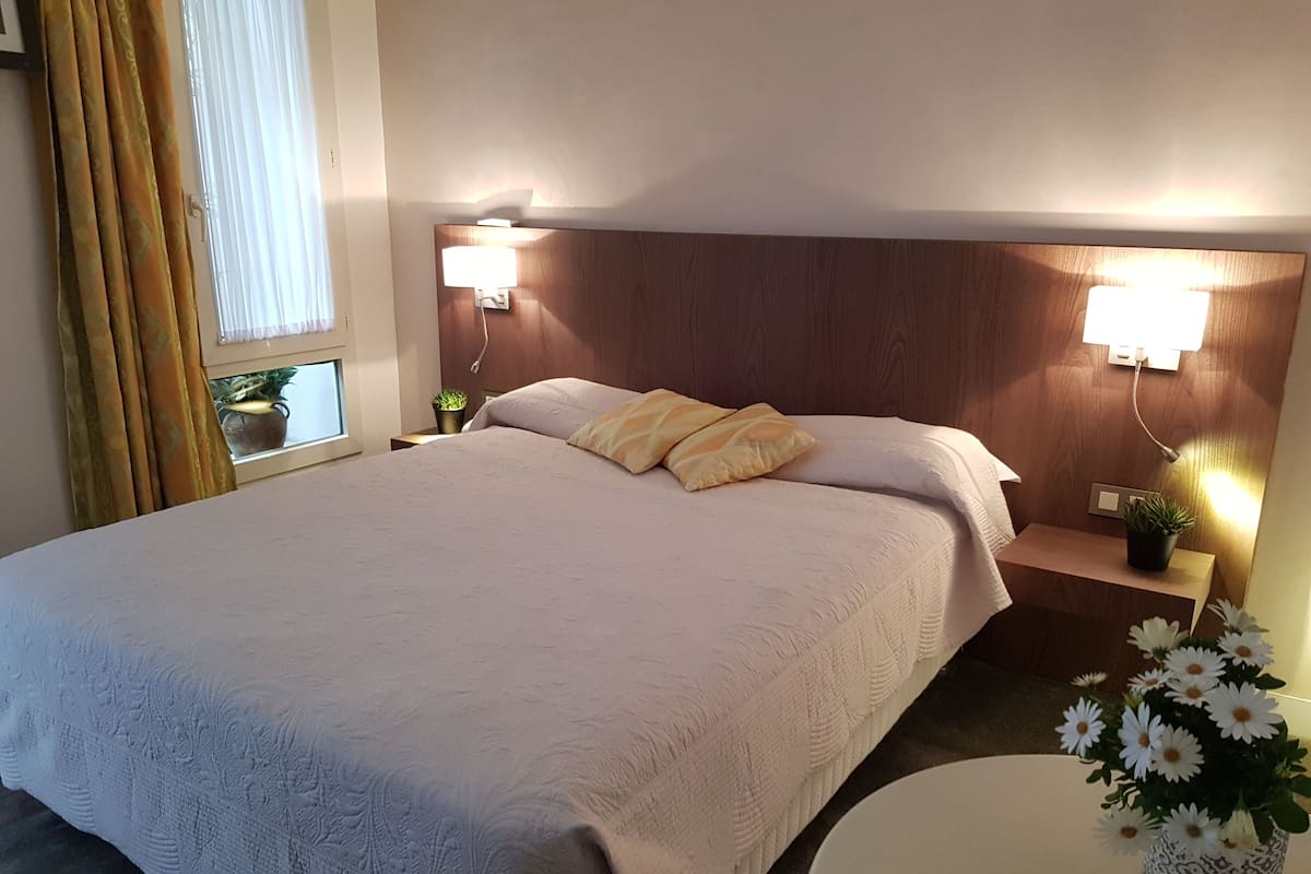 Una cama bien hecha con cabecera, dos mesitas de noche con lámparas, una mesita redonda con un florero y una ventana con cortinas.