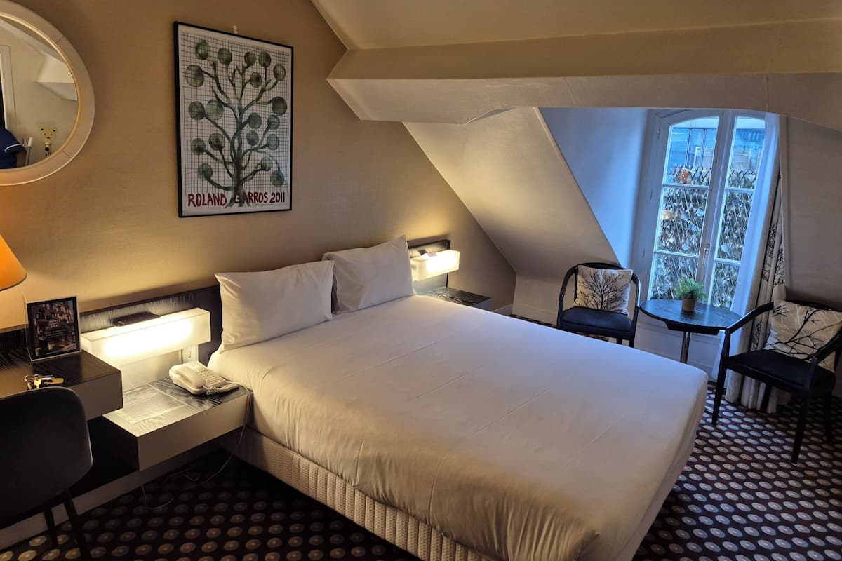 Habitación de hotel con una cama grande, mesita de noche, lámpara, silla y ventana con vistas.