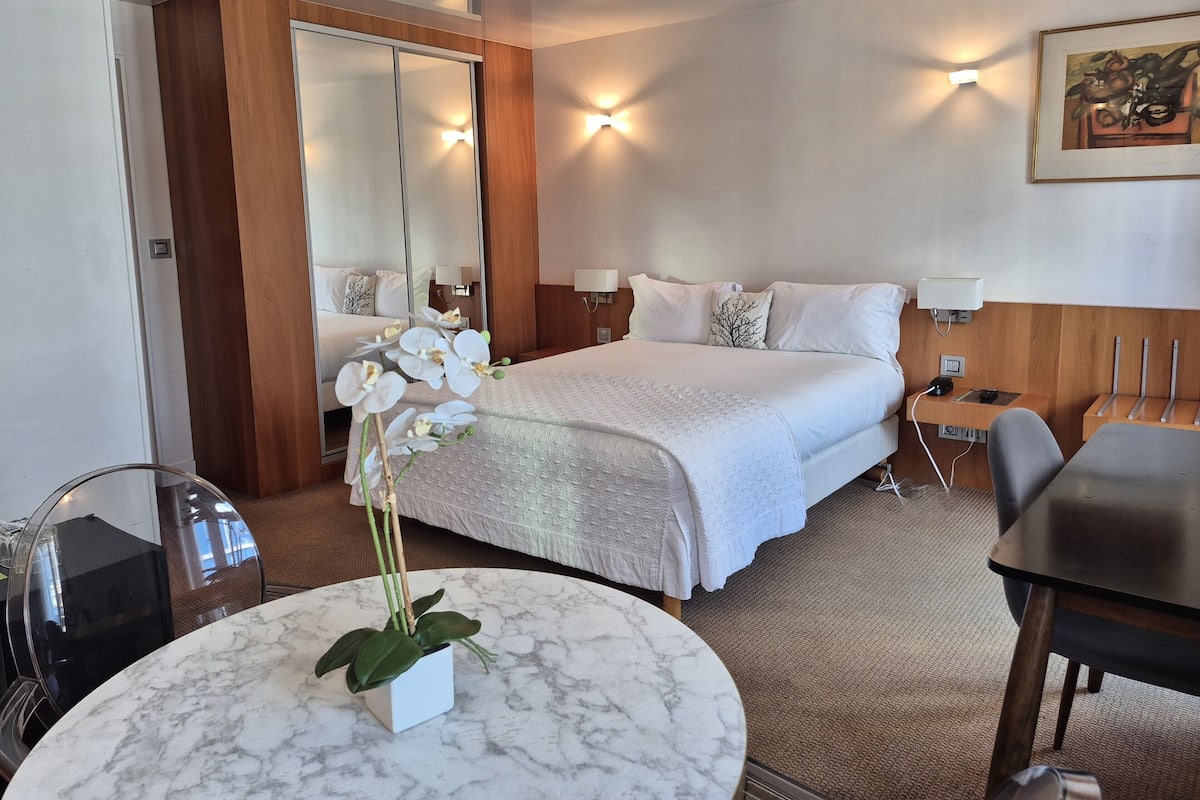 Una habitación de hotel con una cama, mesitas de noche, un escritorio, una silla, una mesa redonda con una planta en maceta y un espejo grande.