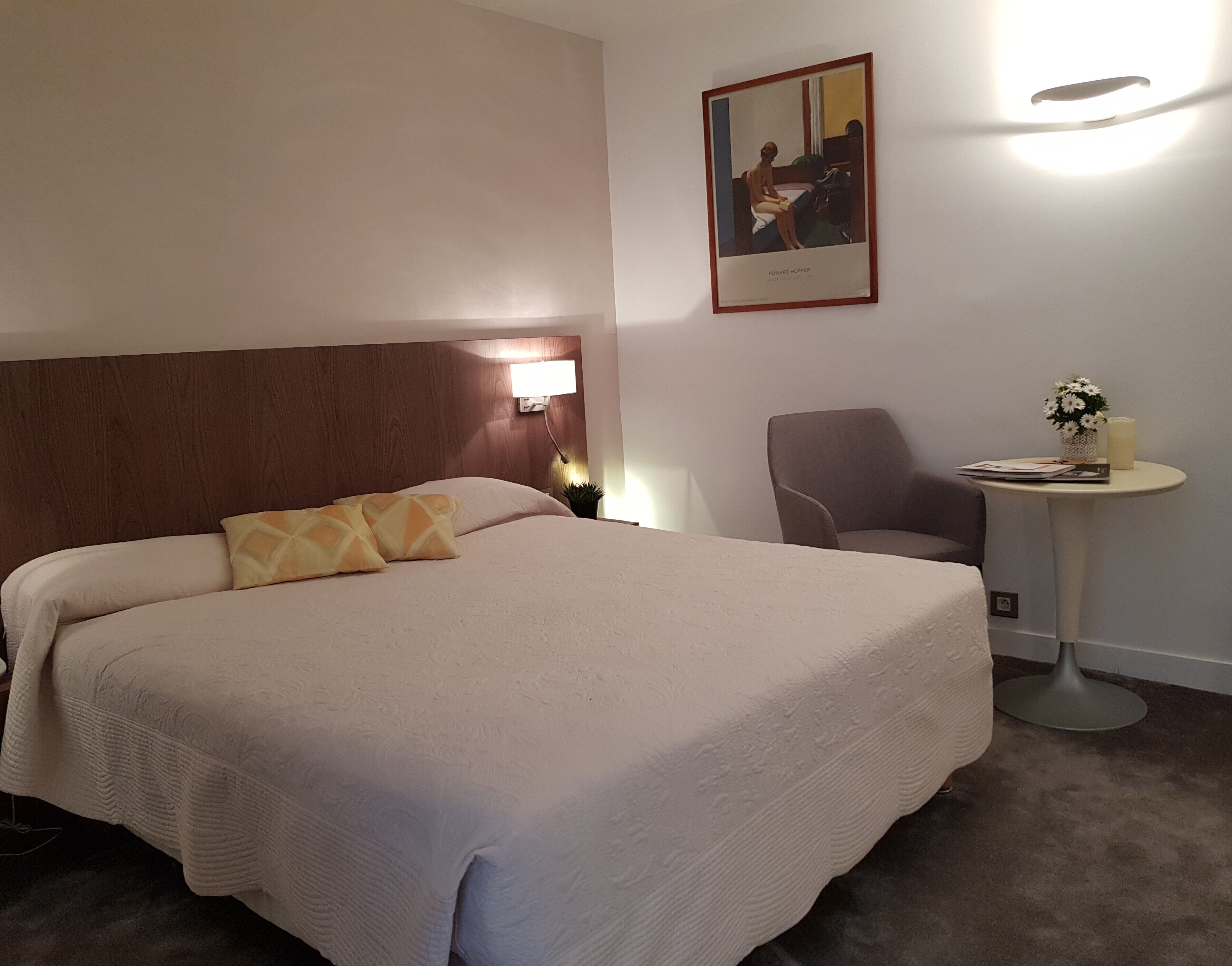 A hotel room with a large bed, a chair, a small table with a vase and candle, and a painting on the wall.