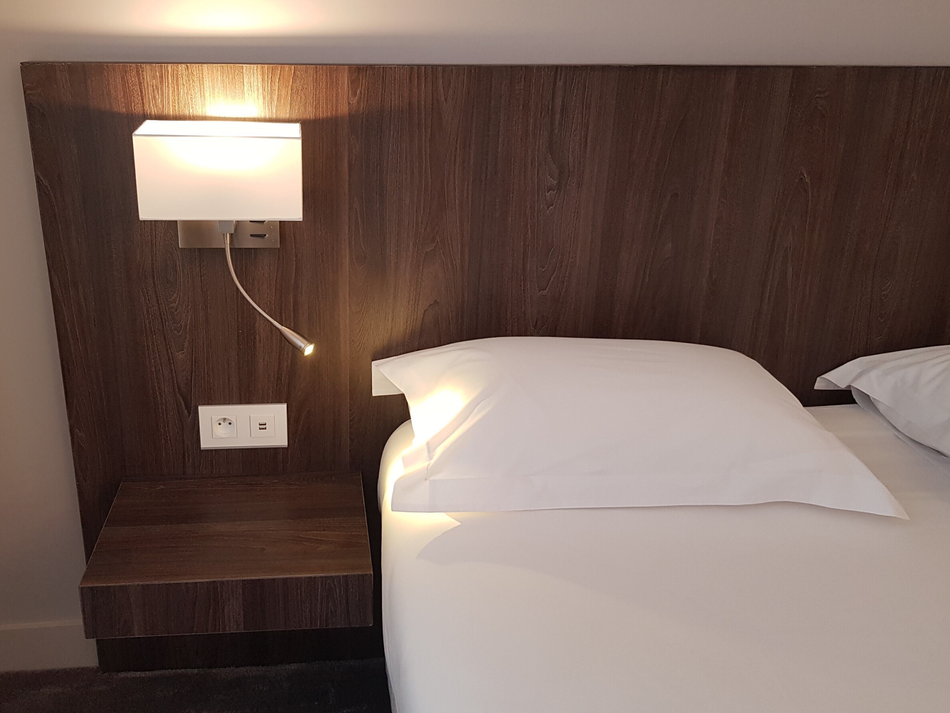 A hotel room with a wooden headboard, a bedside lamp, a power outlet, and a neatly made bed with white linens.