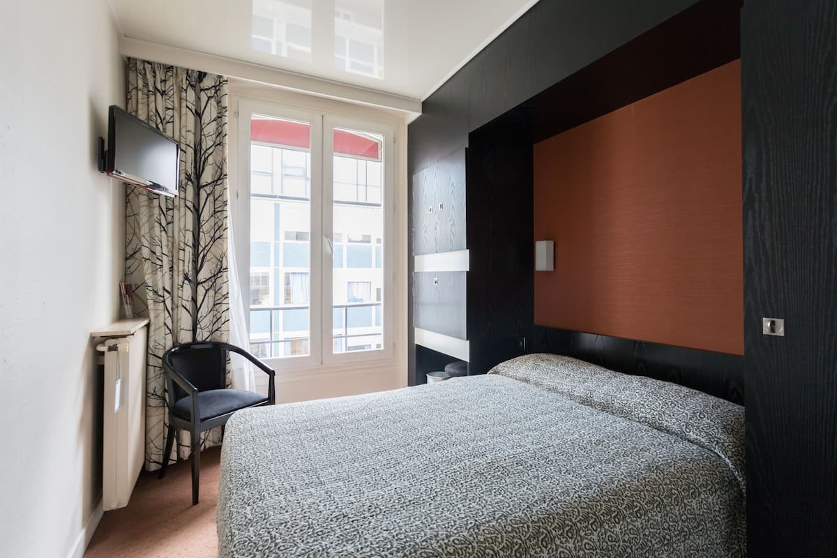 Un dormitorio con cama, una silla, televisor montado en la pared y una ventana con cortinas.