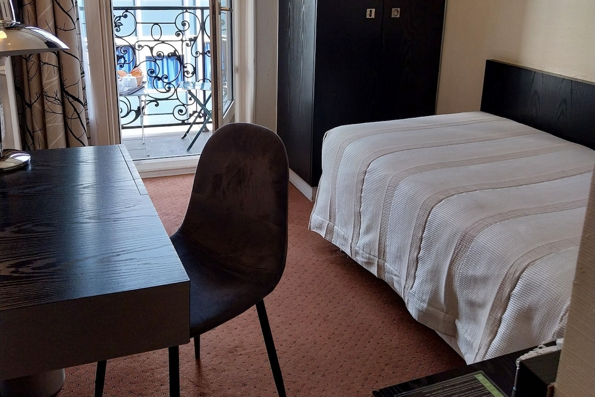 A hotel room with a bed, a desk, a chair, a TV, and a balcony with a view of the city.