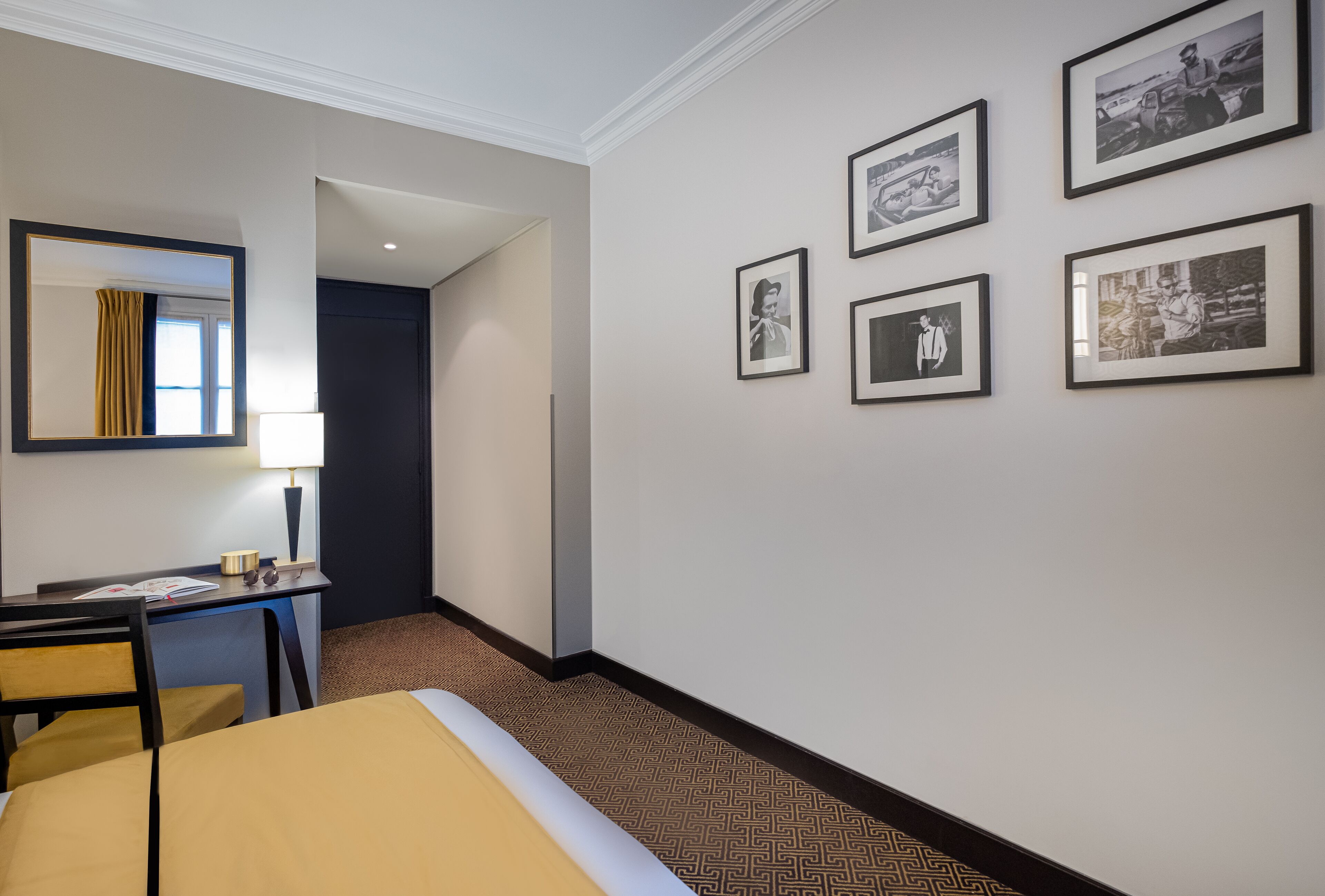 A hotel room with a bed, desk, chair, and framed pictures on the wall.