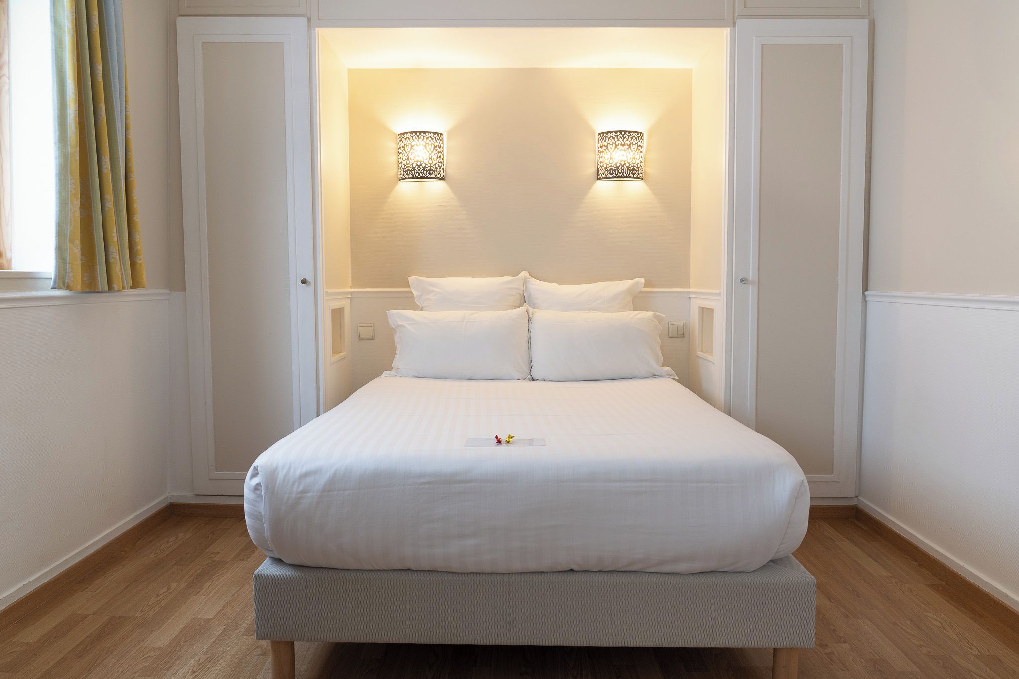 Una cama bien hecha con sábanas blancas, una mesita de noche y dos lámparas fijadas a la pared.
