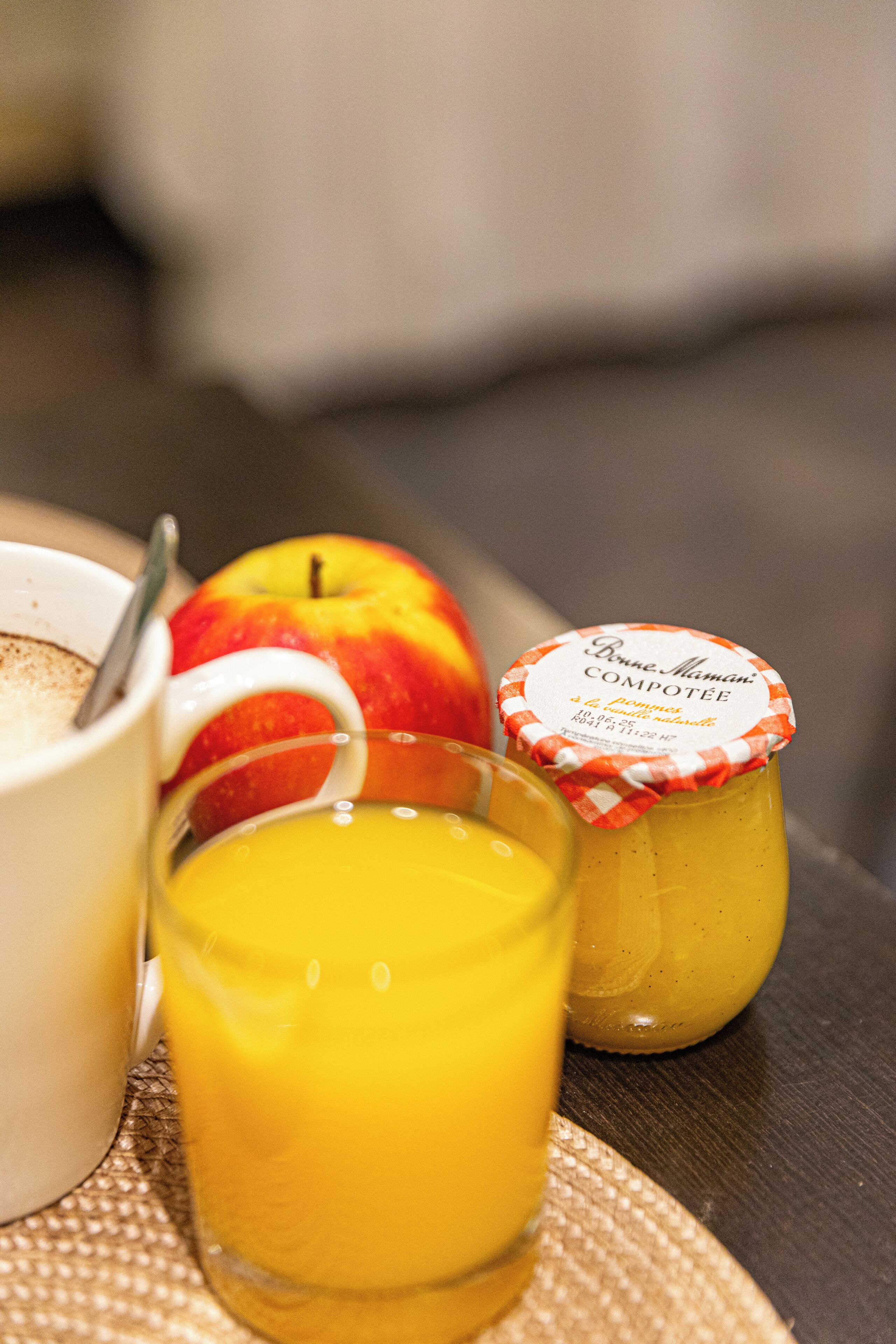 Une table mise pour le petit-déjeuner avec une tasse, une pomme, un verre de jus d’orange et un pot de compote Bonne Maman.