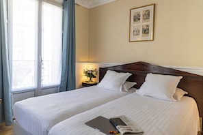 A hotel room with two beds, a nightstand with a lamp and flowers, and a wall with framed pictures. - Hotel Claude Bernard Saint Germain (Paris)