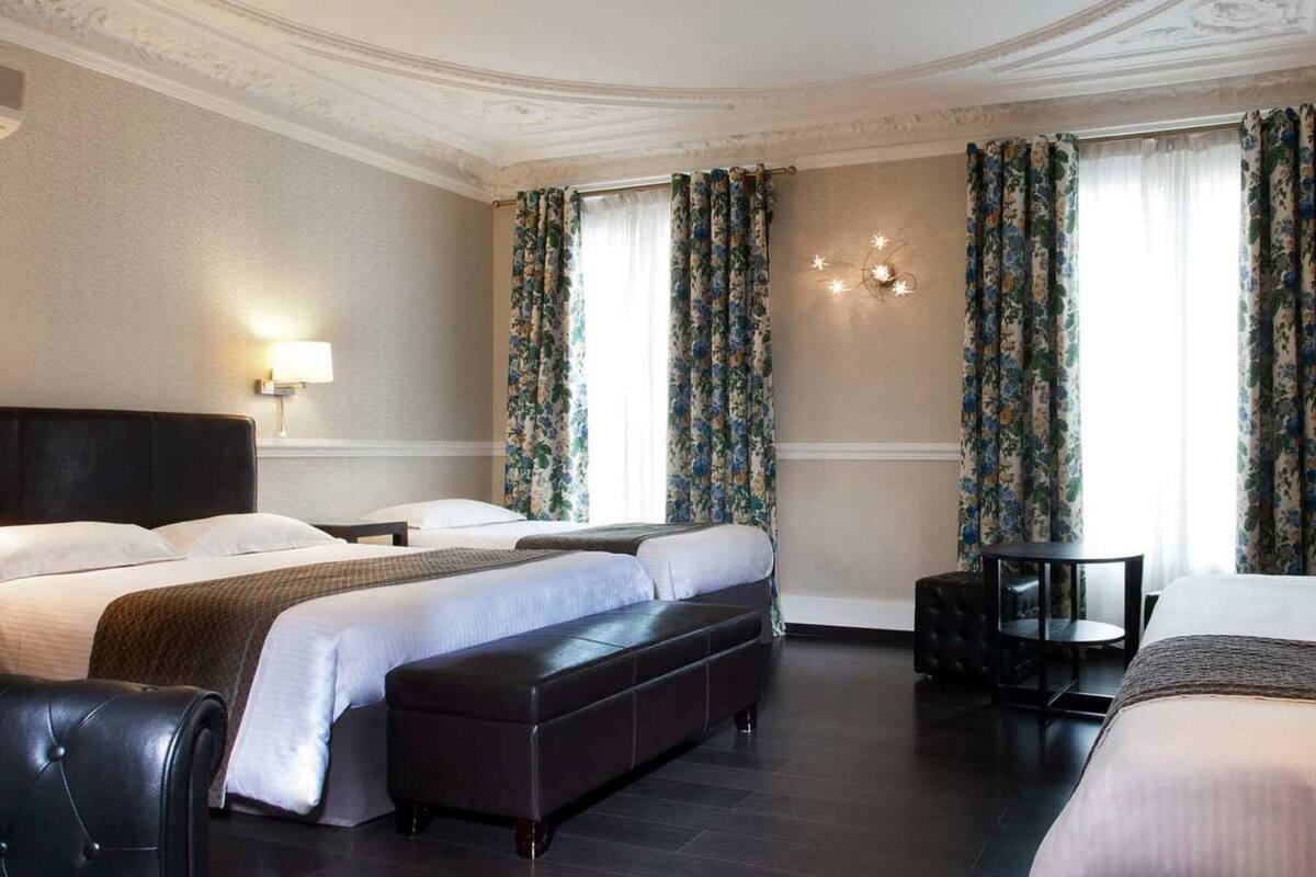 A hotel room with a large bed, a bench, a desk, and a chair. There are two lamps on the nightstands and a chandelier on the wall.