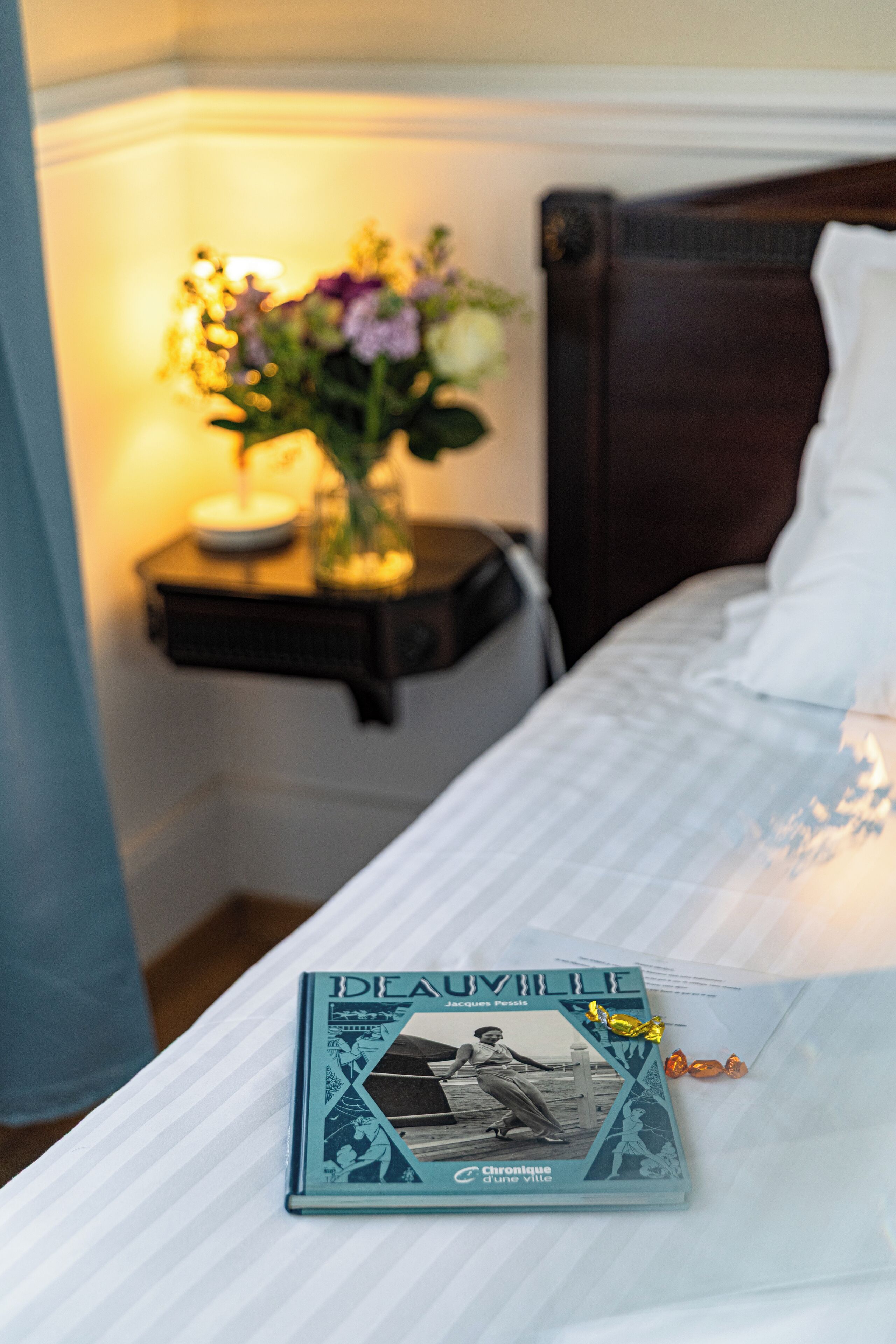 A neatly made bed with a book titled 'DE AUJVILLE' and a small vase with flowers on a bedside table.
